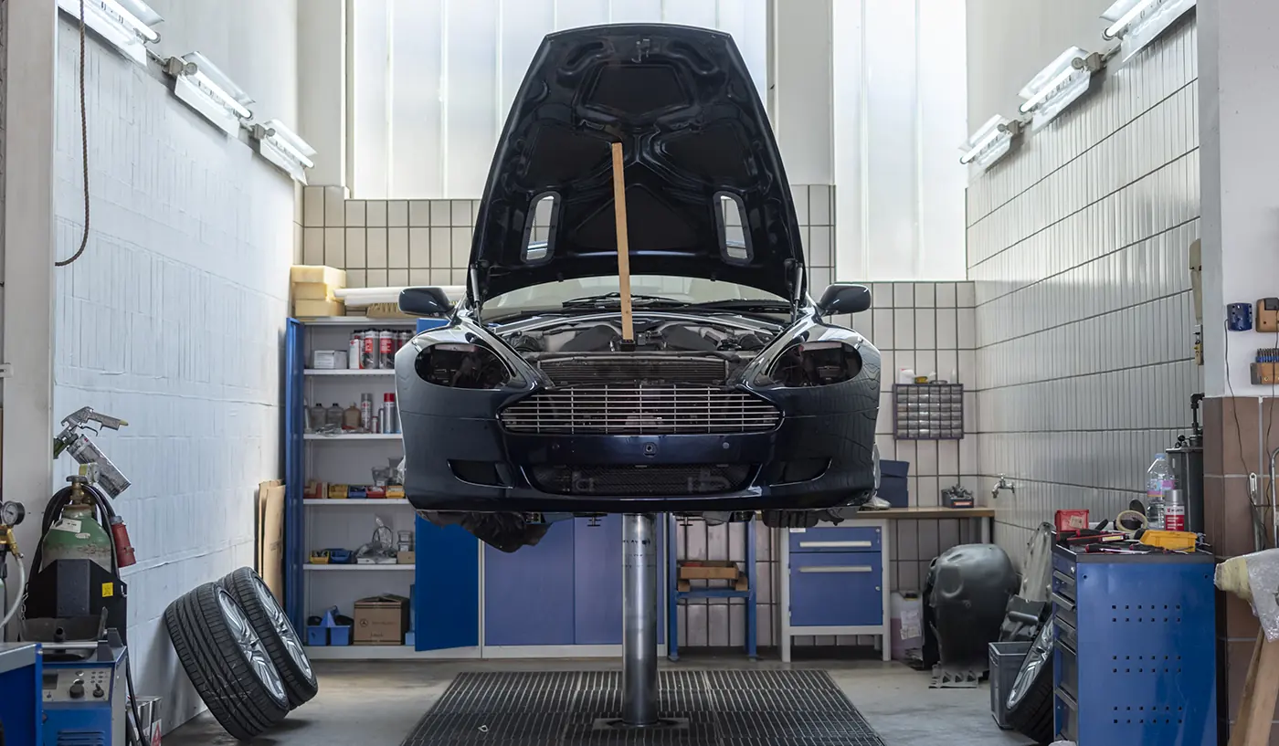 Ein Sportwagen mit geöffneter Motorhaube auf der Hebebühne in der Karosseriewerkstatt, daneben Werkzeugwagen und Regale.