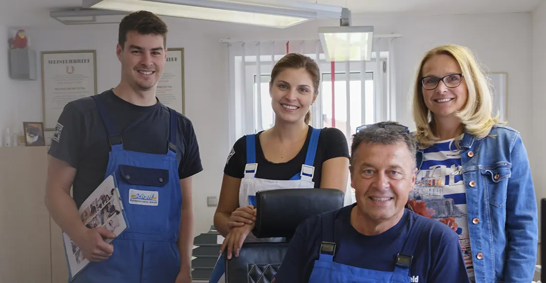 Gerhard Scharf sitzt am Schreibtisch im Büro, hinter ihm stehen drei Mitarbeitende, zwei davon in blauer Arbeitskleidung mit Scharf-Schriftzug.